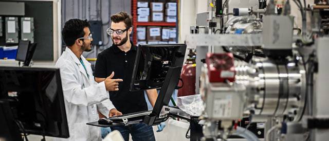 alt = Two male individuals in a manufacturing setting. One is wearing a lab coat and safety glasses pointing at the computer. The other is wearing a black shirt and safety glasses listening to the other individual. There is a variety of systems and equipment in the background.