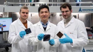 alt = Three male researcher’s wearing white lab coats, blue protective gloves in a laboratory from Angstrom Engineering's partners at EMPA Switzerland present samples of their work, showcasing the newly developed perovskite-perovskite tandem solar cell. An Angstrom Engineering machine is seen in the background with the logo in blue behind the individuals.